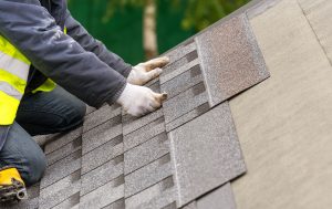 Close up and real photo of professional workman in special protective work wear holding in hands asphalt or bitumen tile and installing it on top of the roof under construction house