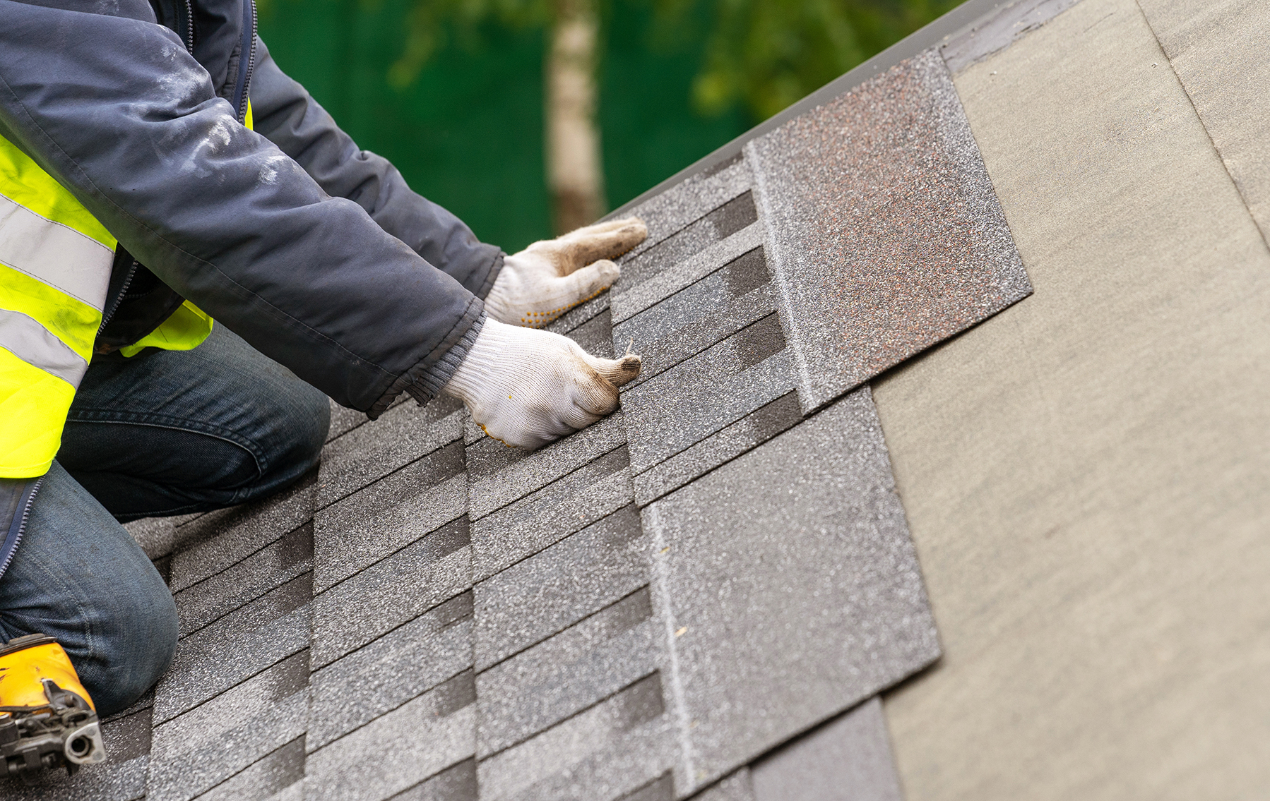 Close up and real photo of professional workman in special protective work wear holding in hands asphalt or bitumen tile and installing it on top of the roof under construction house